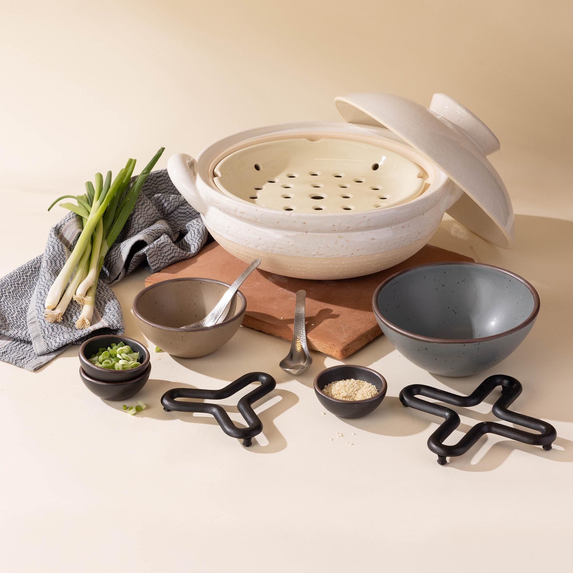 White donabe steamer  arranged with ceramic bowls, and prep ingredients along with cast iron trivets.