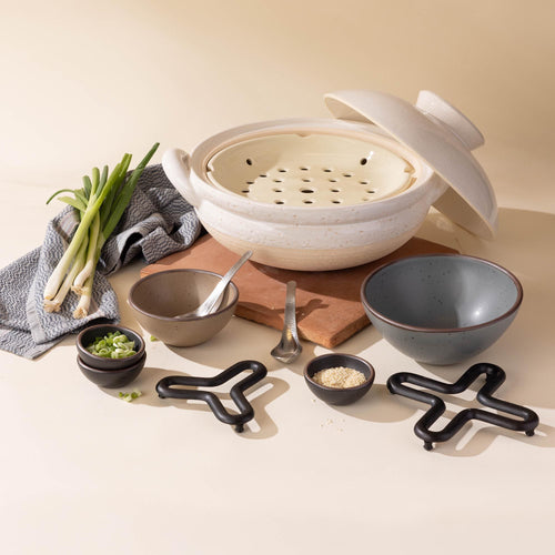 White donabe steamer  arranged with ceramic bowls, and prep ingredients along with cast iron trivets.