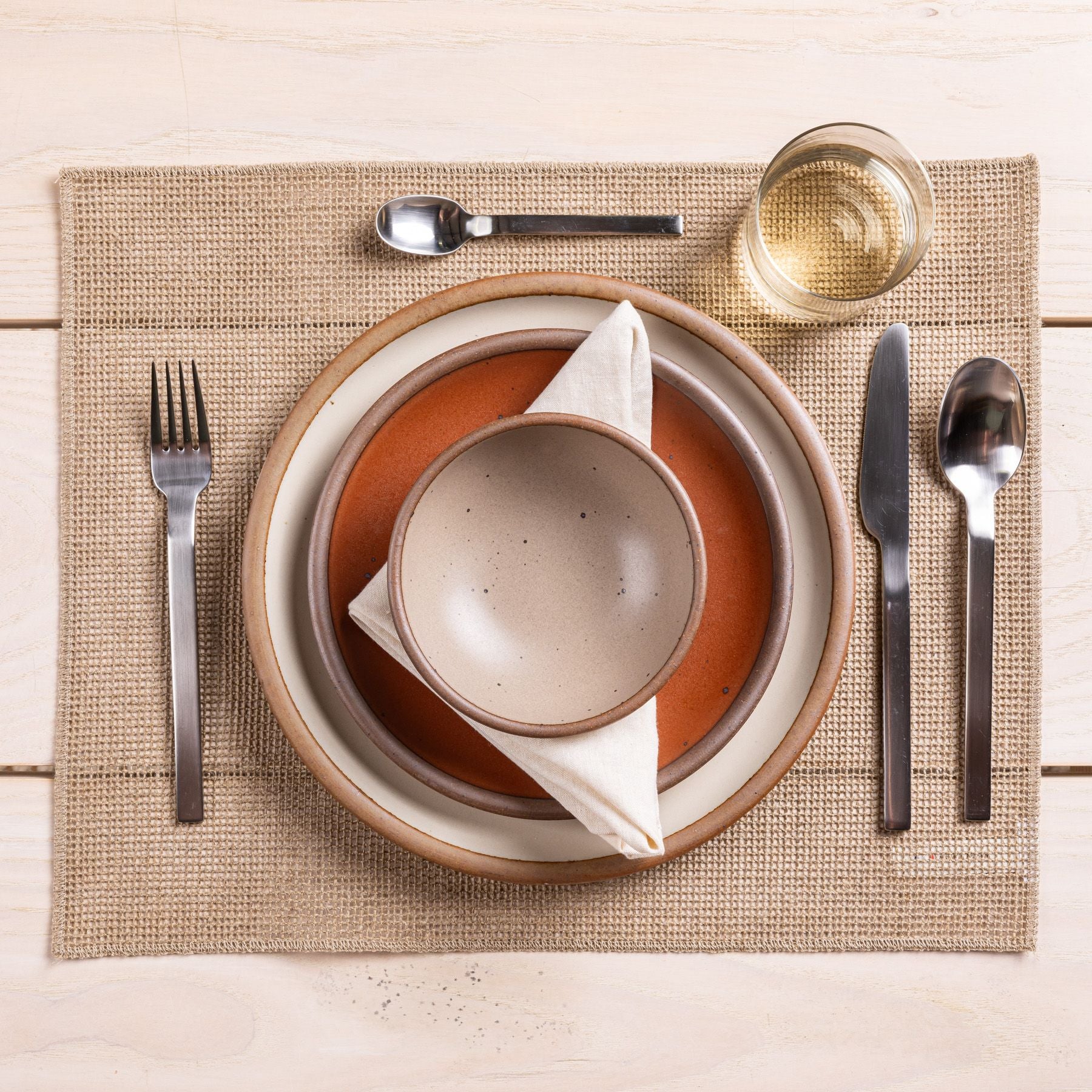 A table setting featuring ceramic plates, a woven placemat, a glass, and steel flatware