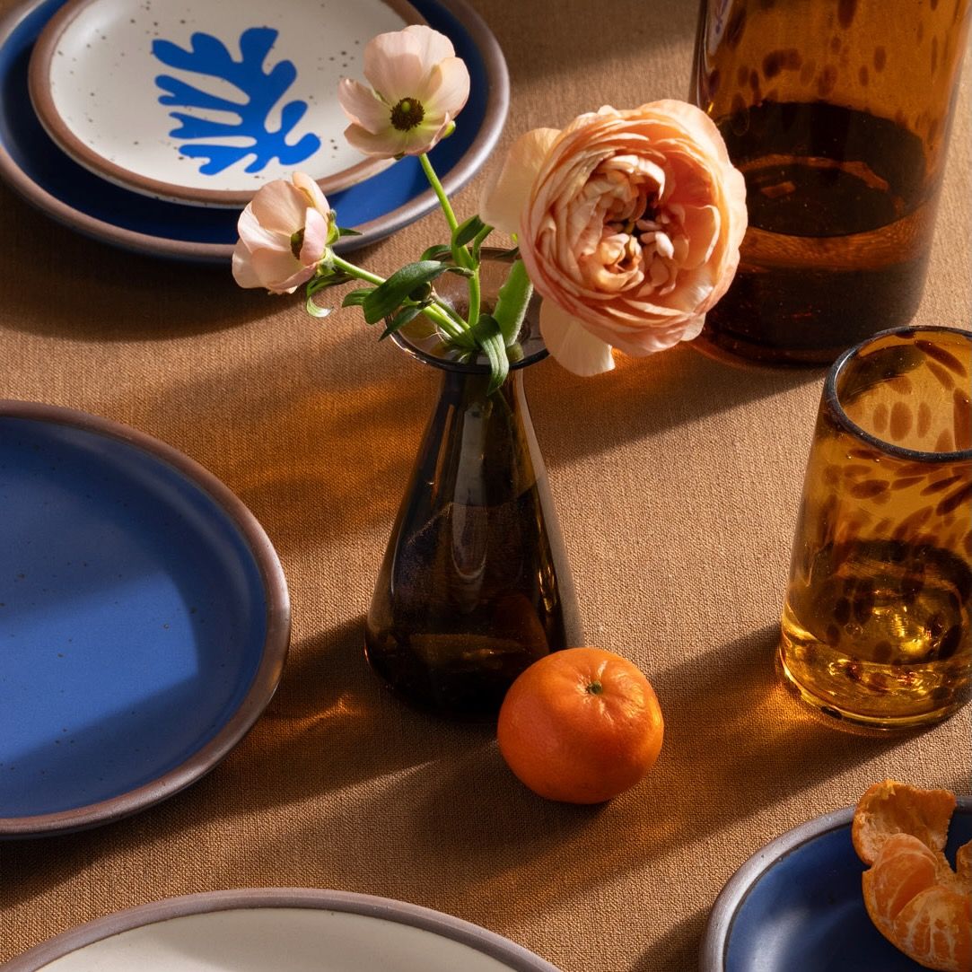 A warm-toned tablescape featuring ceramic dinnerware in blue and cream sits alongside amber glassware, a brown glass vase with soft peach flowers, and a clementine for a pop of color.