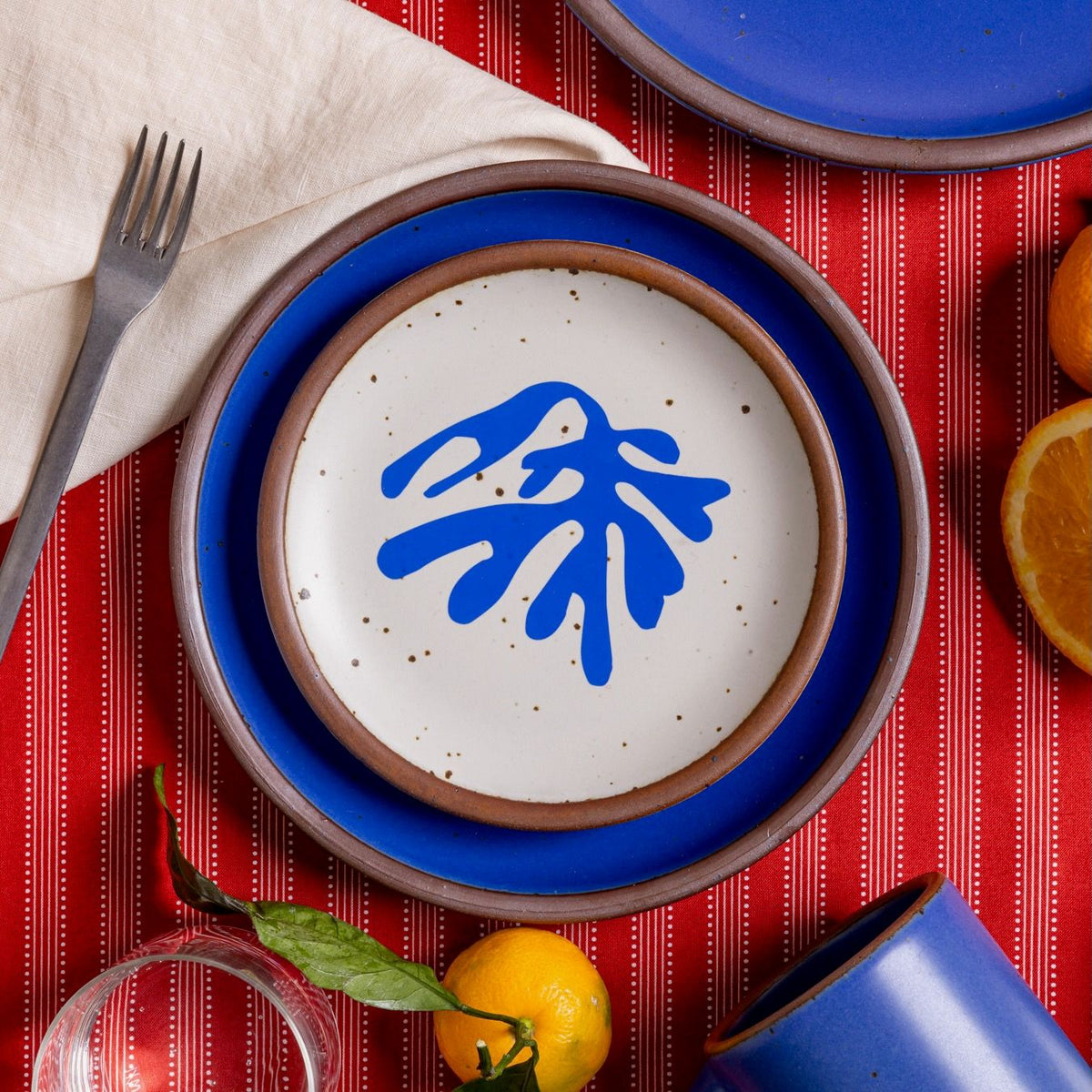 A stack of plates including one that has a blue frond-like cut-out layered with a ceramic plate in a cobalt blue color on a red tablecloth with oranges, a fork, and glass.