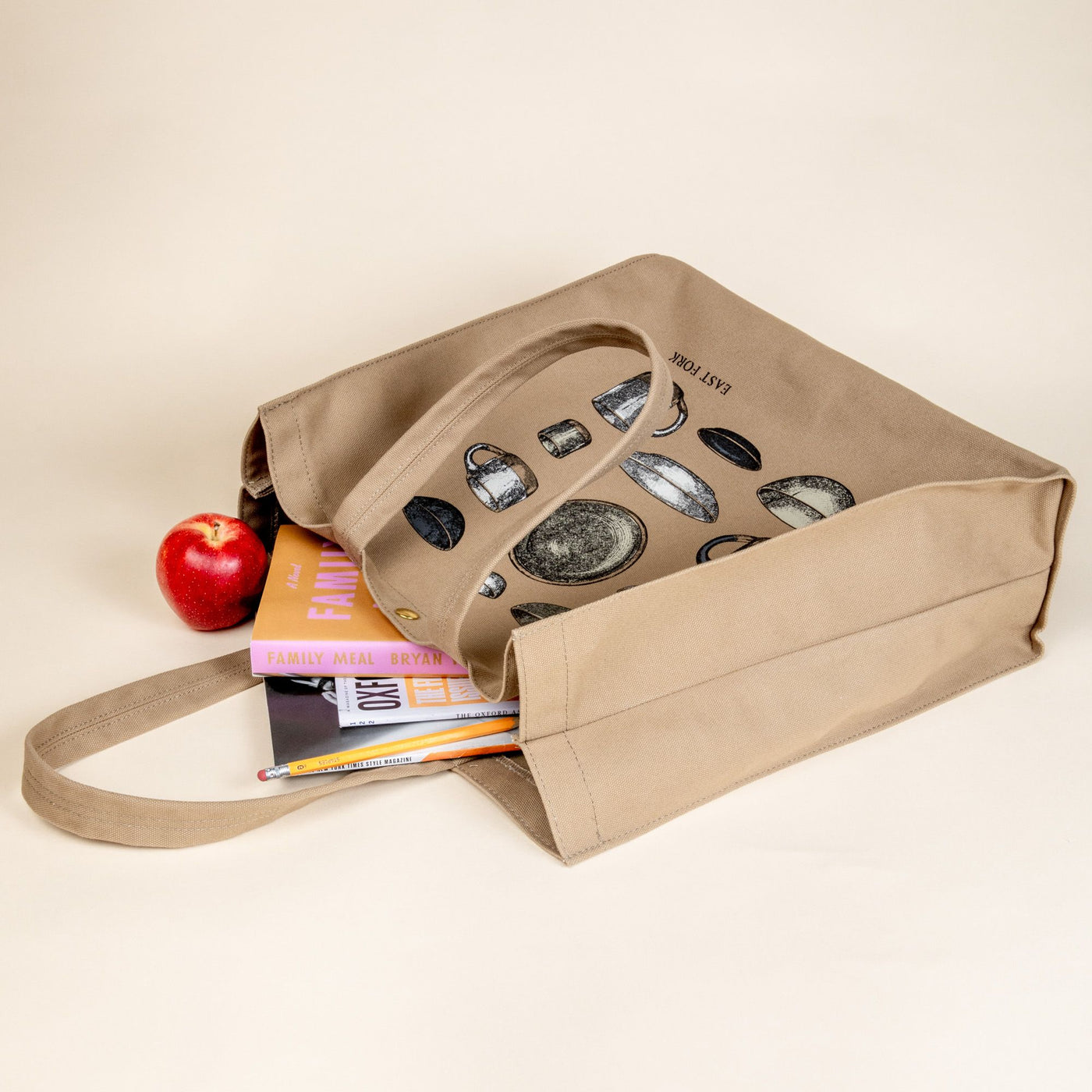 A sturdy canvas tan tote bag on its side with handles and a metal closure button at the top. On the bag is a printed illustration in neutral colors of bowls, plates, and mugs. Books, a pencil, and an apple are falling out of the bag.