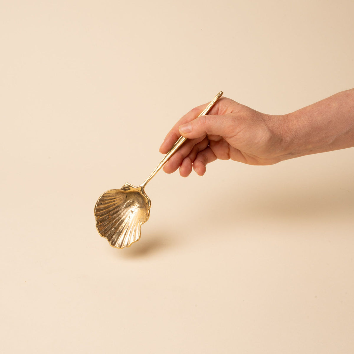 Hand holds out a brass spoon with a scalloped shell-shaped bowl and long handle, set against a neutral beige background.