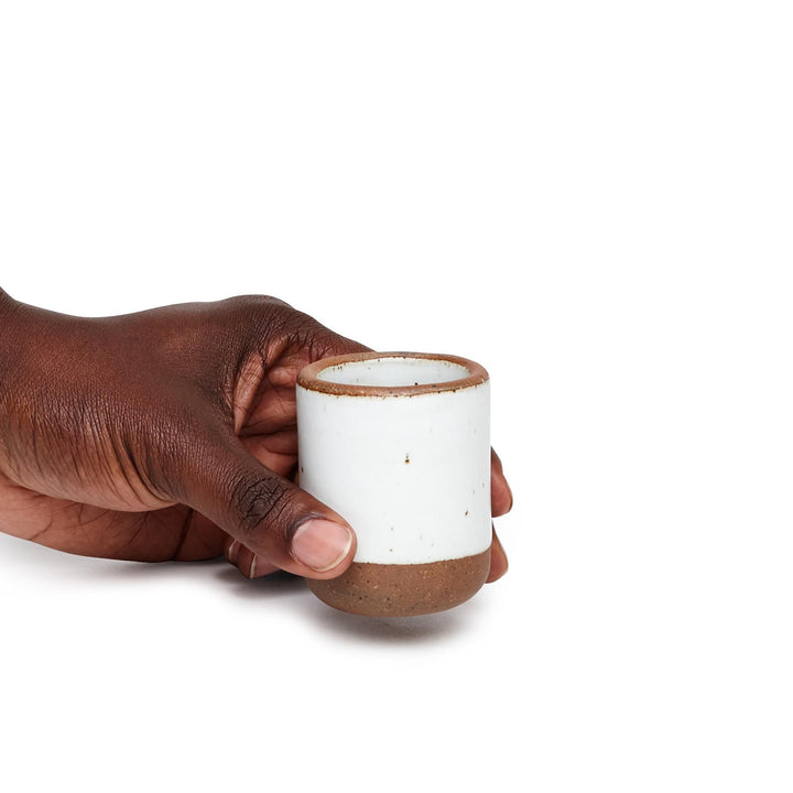 A hand holding a small, short ceramic mug cup in a cool white color featuring iron speckles and unglazed rim and bottom base.