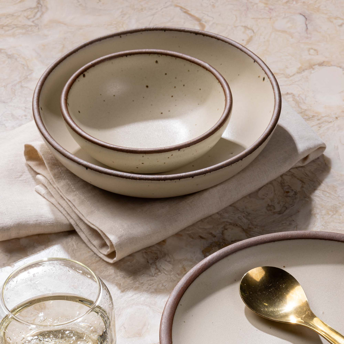 Two nesting ceramic bowls in an off-white color and sitting on a folded napkin. Off to the side is a side plate with a brass spoon.