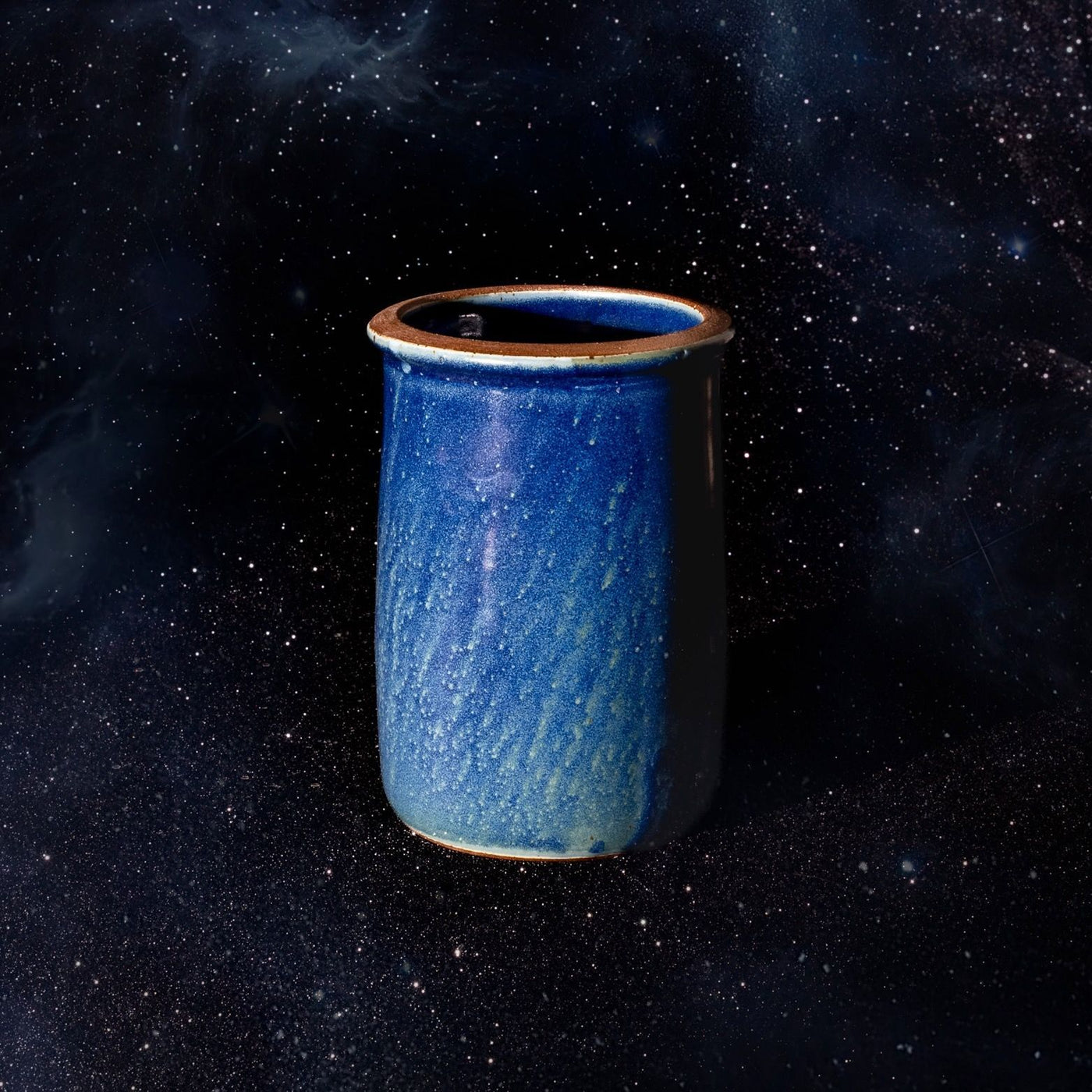 A cylindrical ceramic utensil crock in a reflective dappled deep blue color featuring an unglazed rim, against a dark starry background