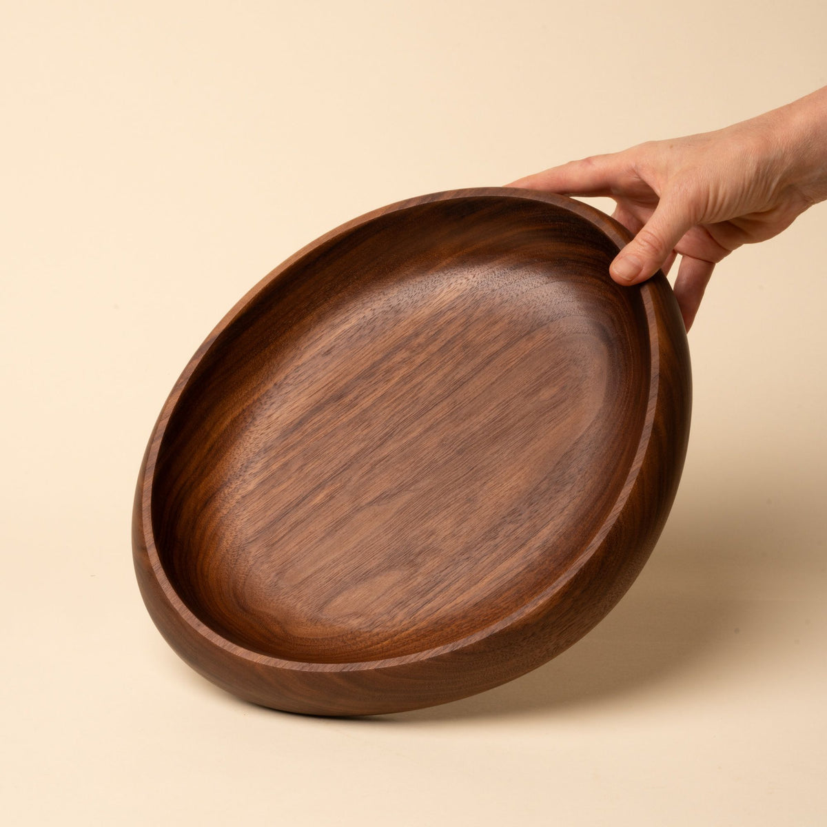 Hand holds out a shallow carved round walnut wood dish with smooth curved edges, resting on a beige background.