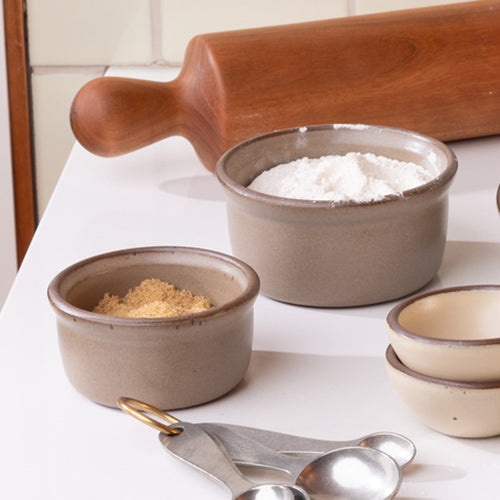 A small and medium ceramic ramekins with flour and sugar, on a countertop with baking accessories.