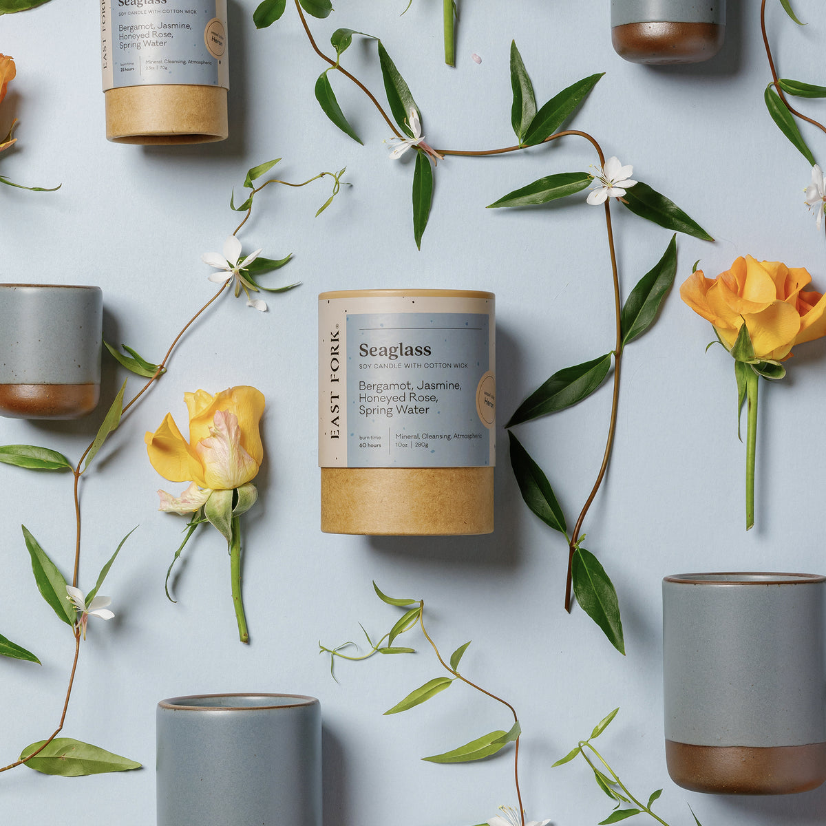 An artful layout of The Candle in Seaglass - centered is a cardboard packaging tube with the candle's name, surrounded by small and large ceramic candles in a cylindrical vessel in a blue-grey color, surrounded by jasmine, roses, and bergamot.