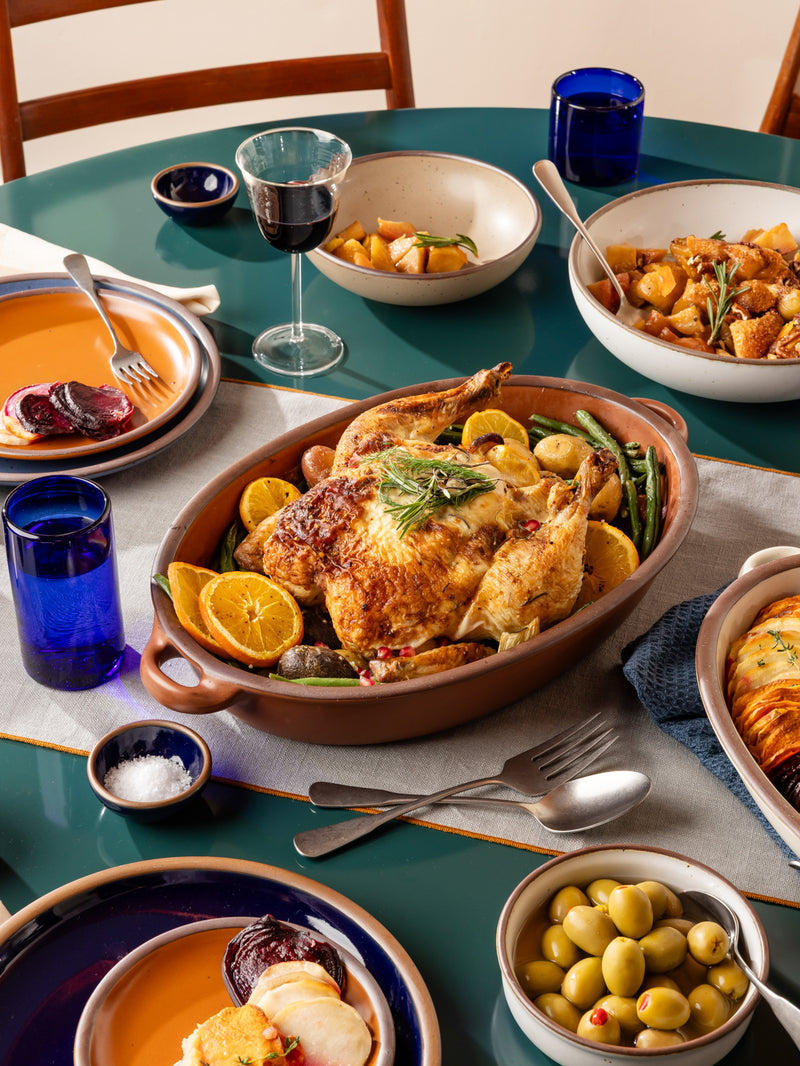 A roasted whole chicken in a cool terracotta ceramic oval baker dish sits on a teal dining table, surrounded by side dishes, blue glass cups, a glass of red wine, and utensils in a styled home dining setting.
