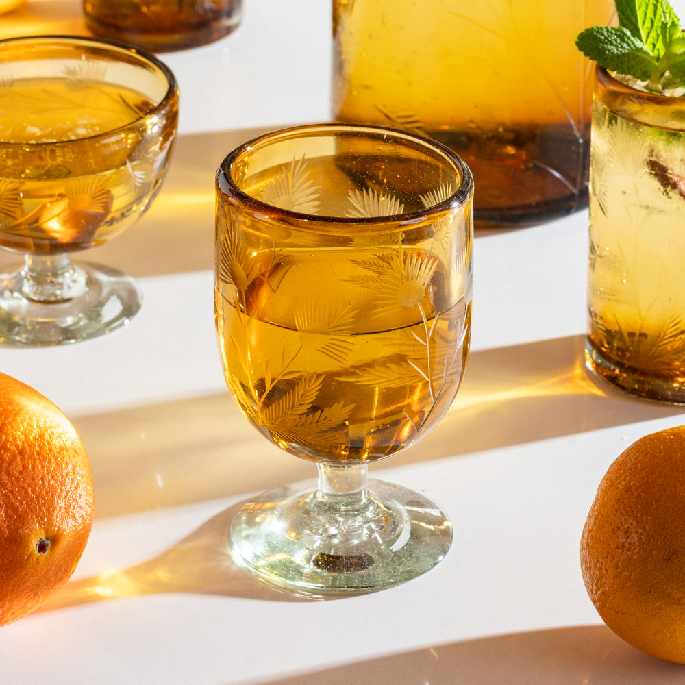 Amber etched stemmed glass filled with a clear beverage beside amatching glassware and oranges.