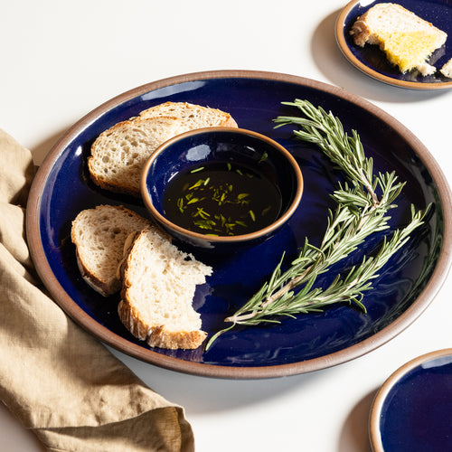 A dark glossy blue ceramic serving platter with a brown rim, styled with sliced bread, a small bowl of oil, and rosemary sprigs on a white surface.