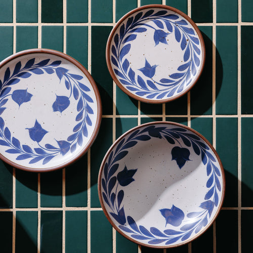An overhead view of a dinner plate, side plate, and shallow bowl with a hand-painted blue leaf and flower border on a white background with an exposed clay rim, against a teal tile background.
