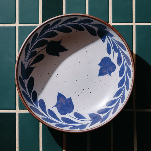 An overhead view of a shallow bowl with a hand-painted blue leaf and flower border on a white background with an exposed clay rim, against a teal tile background.