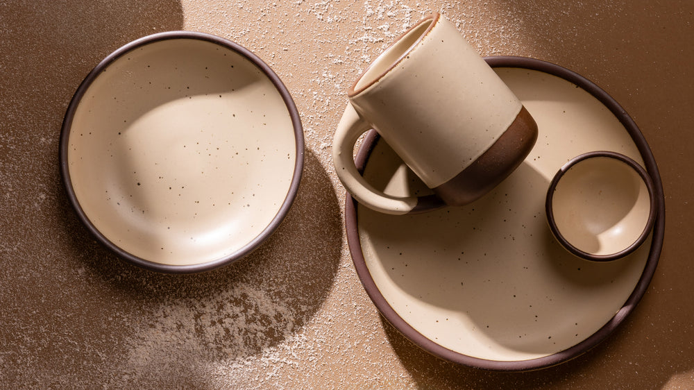 A set of handmade ceramic tableware in a warm cream color with unglazed base including a bowl, a mug resting on a plate, and a small bowl, arranged on a textured brown surface.