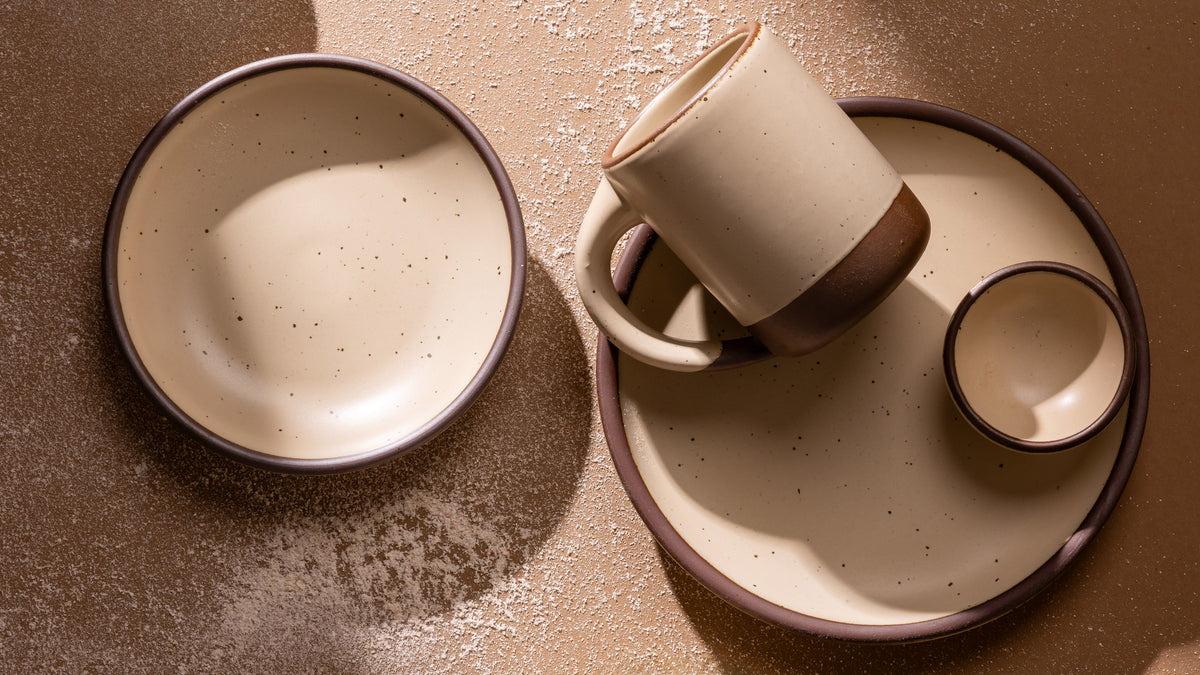 A set of handmade ceramic tableware in a warm cream color with unglazed base including a bowl, a mug resting on a plate, and a small bowl, arranged on a textured brown surface.