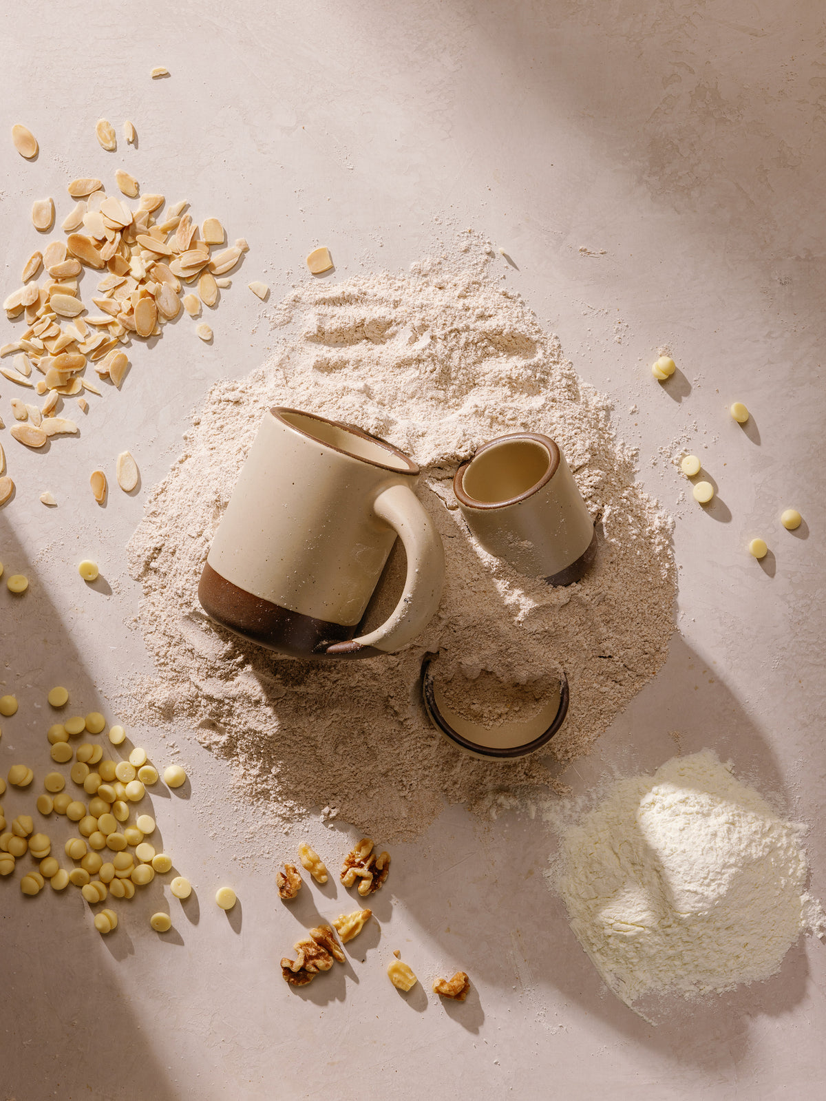 Two ceramic mugs, one larger cream mug with an unglazed base and one smaller matching beige mug, arranged on a mound of flour with scattered oats, walnuts, and a small bowl of flour on a warm beige surface.