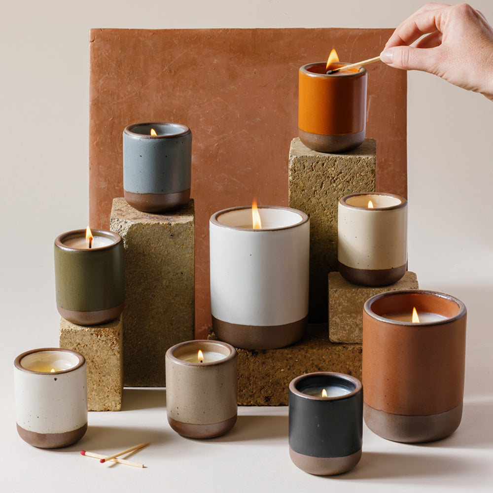 Small and large candles in ceramic vessels in a variety of colors artfully arranged, all lit, and a hand lighting one at the top.