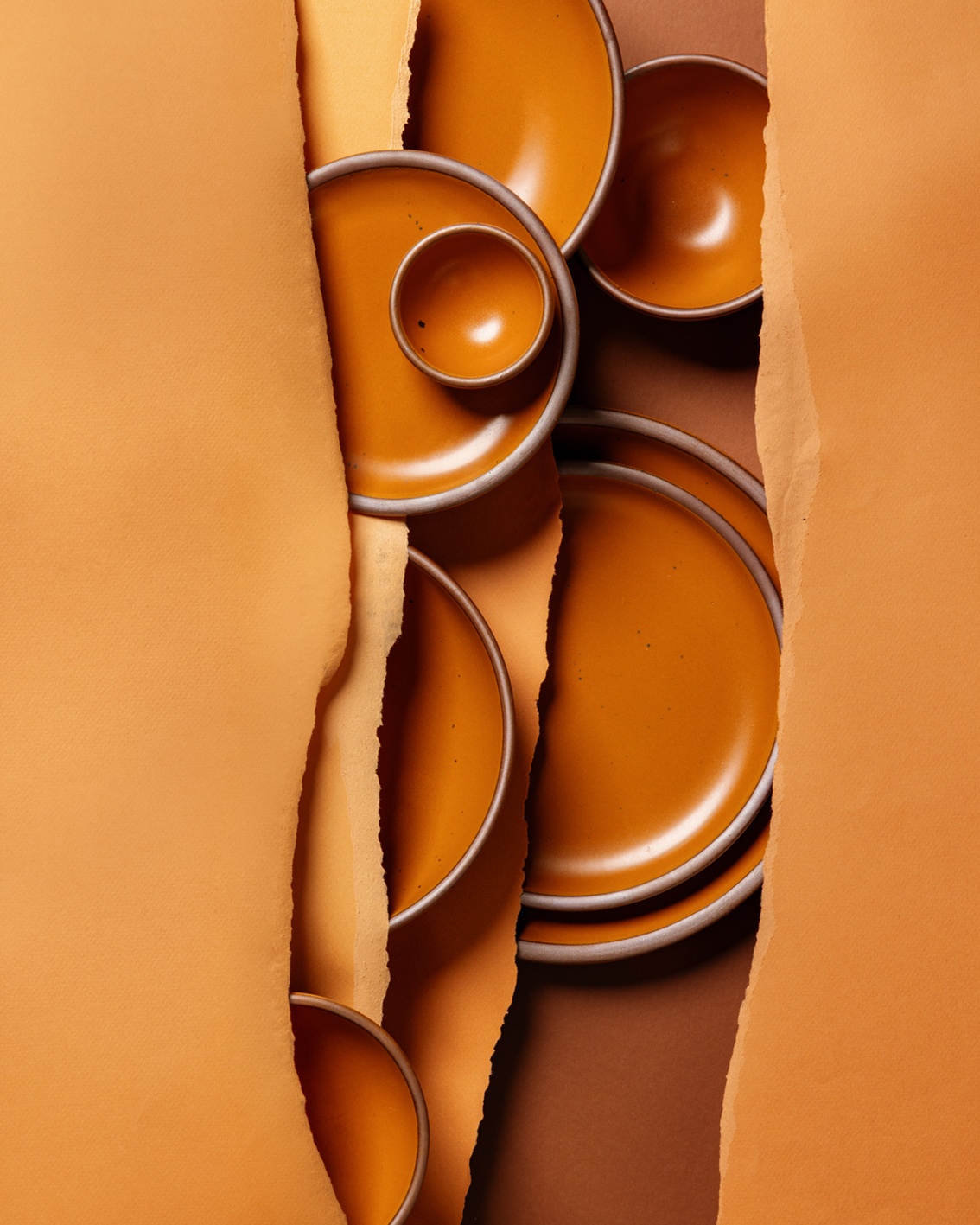A styled flat-lay of earthy orange colored ceramic plates and bowls, partially obscured by torn orange paper on both sides, set against a warm brown background.