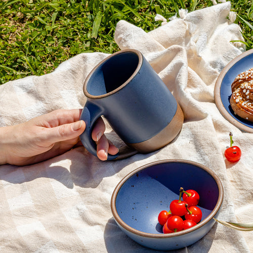 A hand holds a big ceramic mug in a cool blue color with matching dishes on a picnic blanket outside.