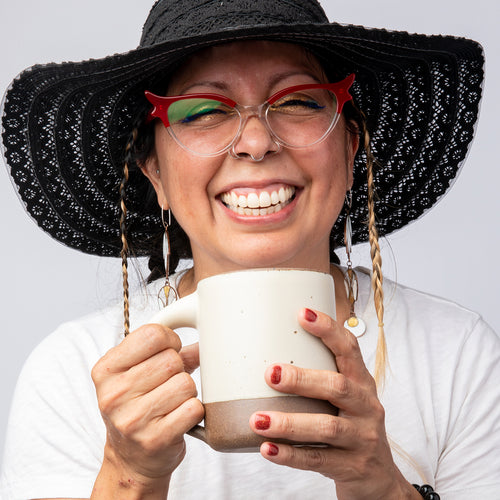 A person smiling wearing a big hat and holding a large ceramic mug in a warm off-white color with an unglazed rim.