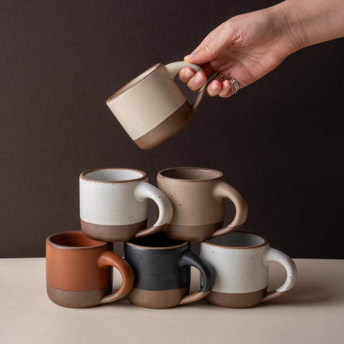 Six short ceramic mugs in assorted neutral, cool terracotta, and black glazes, with a hand holding one mug above the stack.