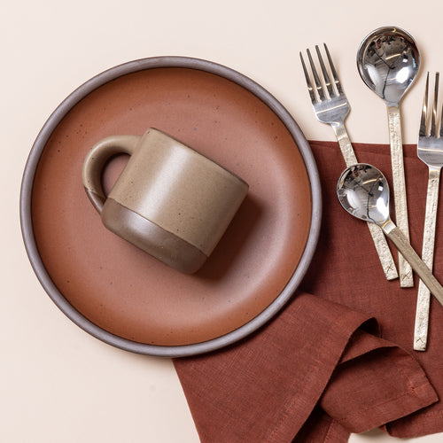 Short ceramic mug in Morel resting on a cool terracotta plate beside silver flatware and a rust-colored napkin.