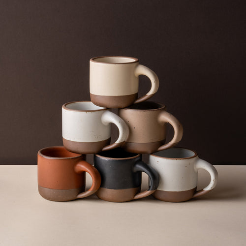 Six short ceramic mugs in assorted neutral, cool terracotta, and black glazes.