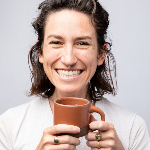 Connie holding a small ceramic mug in a cool terracotta color and smiling big.