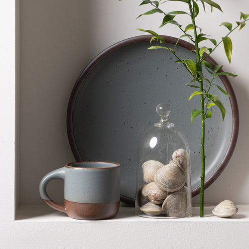 Gray-blue ceramic short mug beside a large matching plate, glass cloche filled with shells, and a green branch.