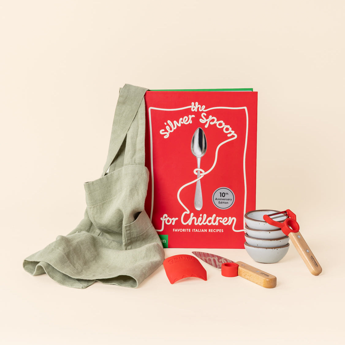 A children's cooking set featuring The Silver Spoon for Children cookbook, a sage green apron, four small ceramic bowls in a matte cool white color with unglazed rims, arranged against a cream background.