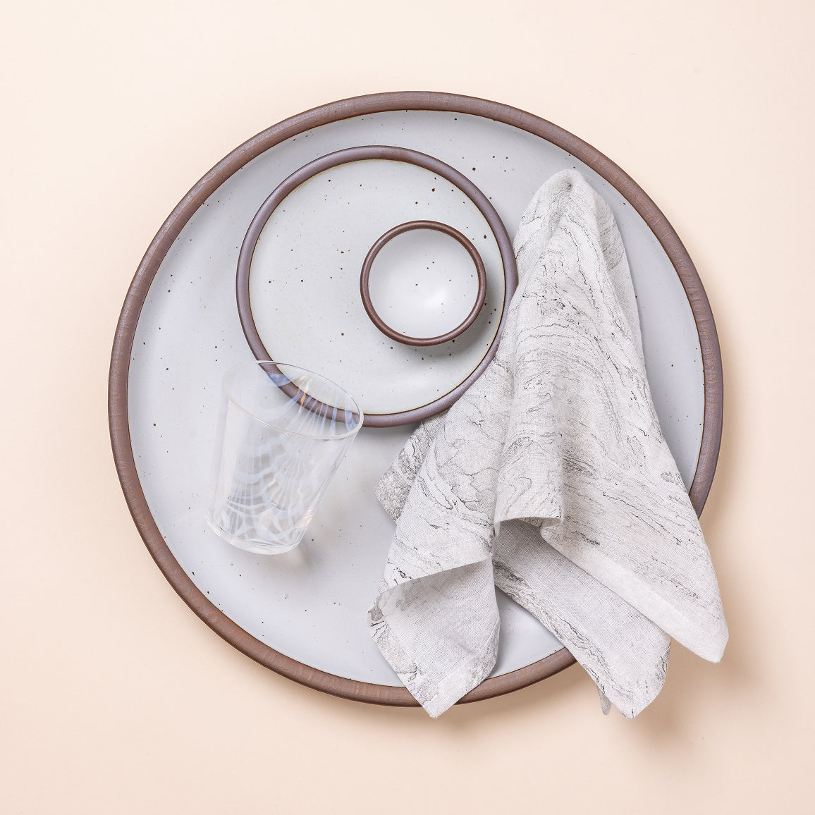 Large white speckled ceramic platter with an unglazed rim, matching side plate and small bowl, etched drinking glass, and patterned linen napkin.
