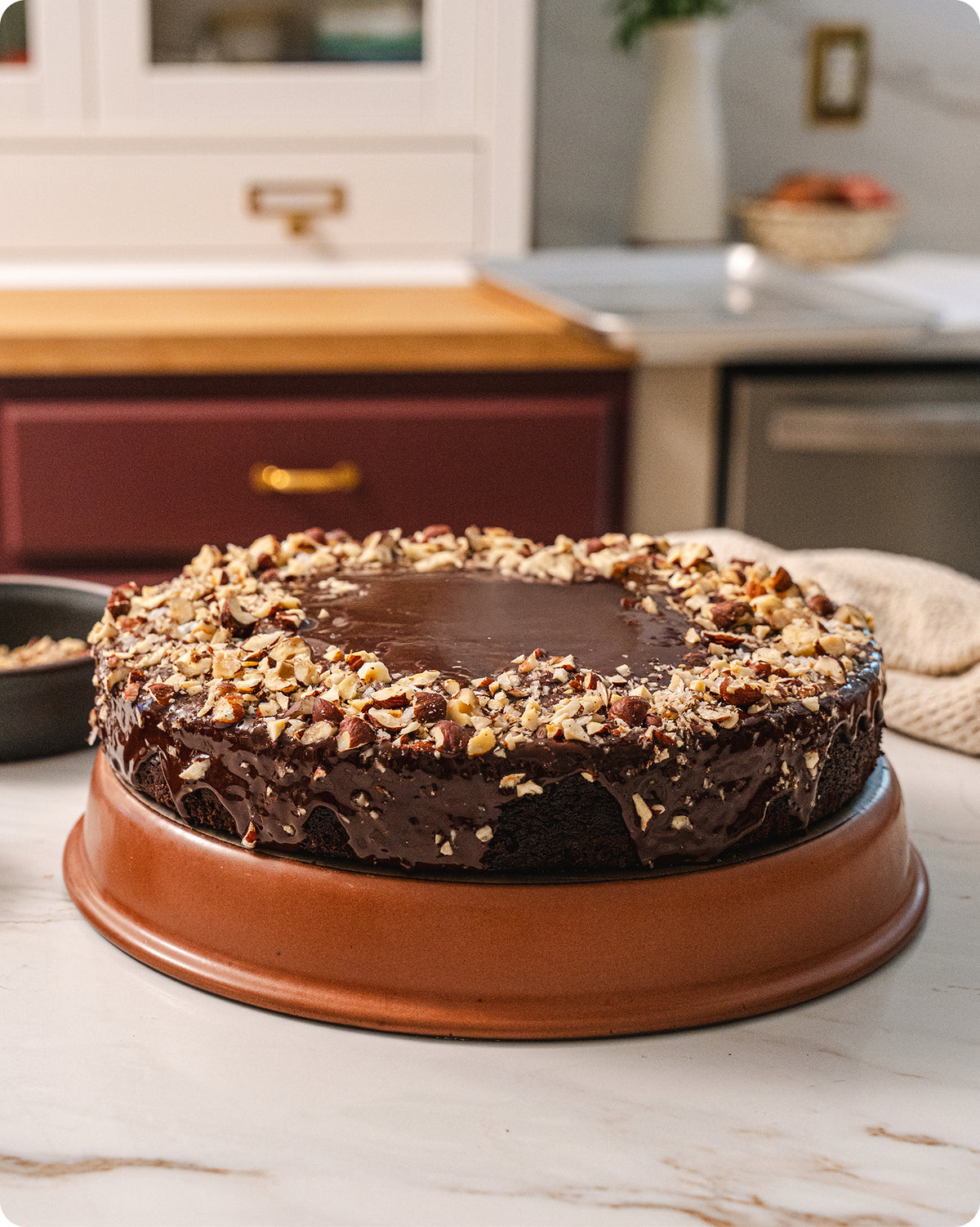 A round chocolate cake with glossy chocolate frosting and chopped nuts on top, displayed on a round terracotta-colored shallow baker in a kitchen setting.