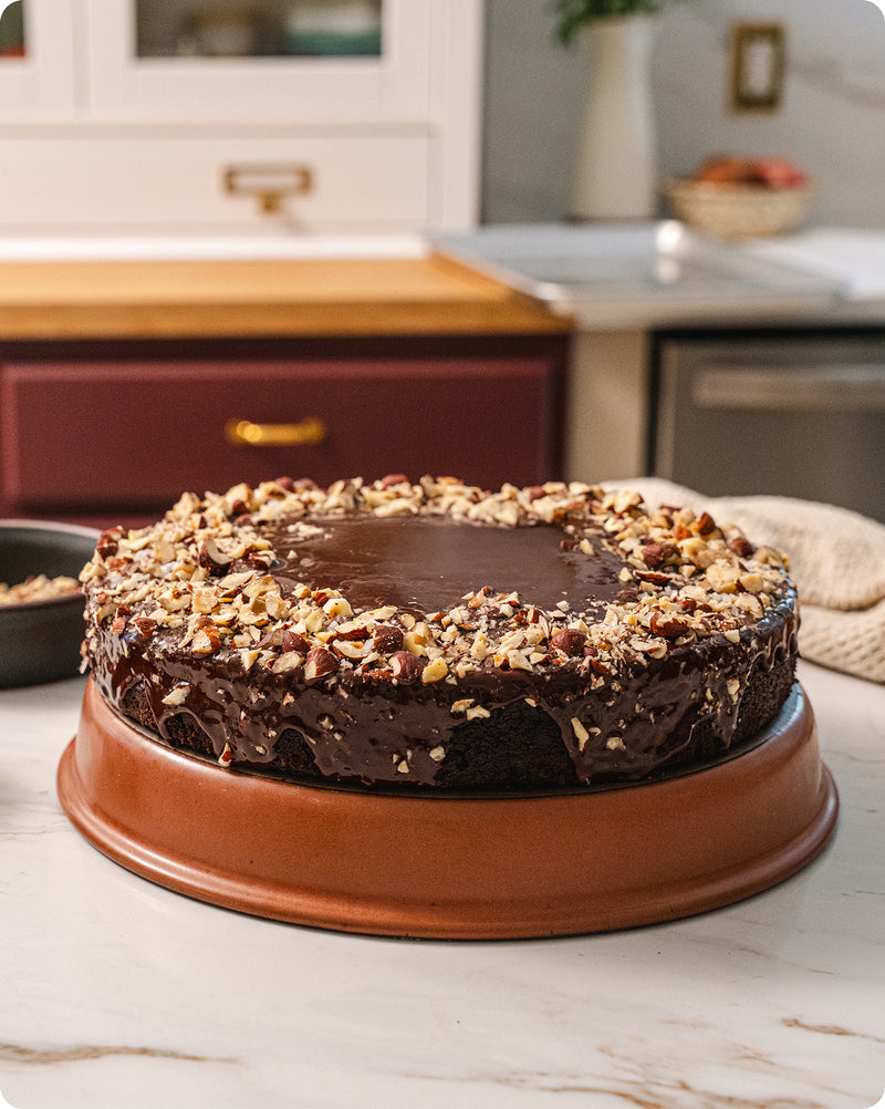 A round chocolate cake with glossy chocolate frosting and chopped nuts on top, displayed on a round terracotta-colored shallow baker in a kitchen setting.
