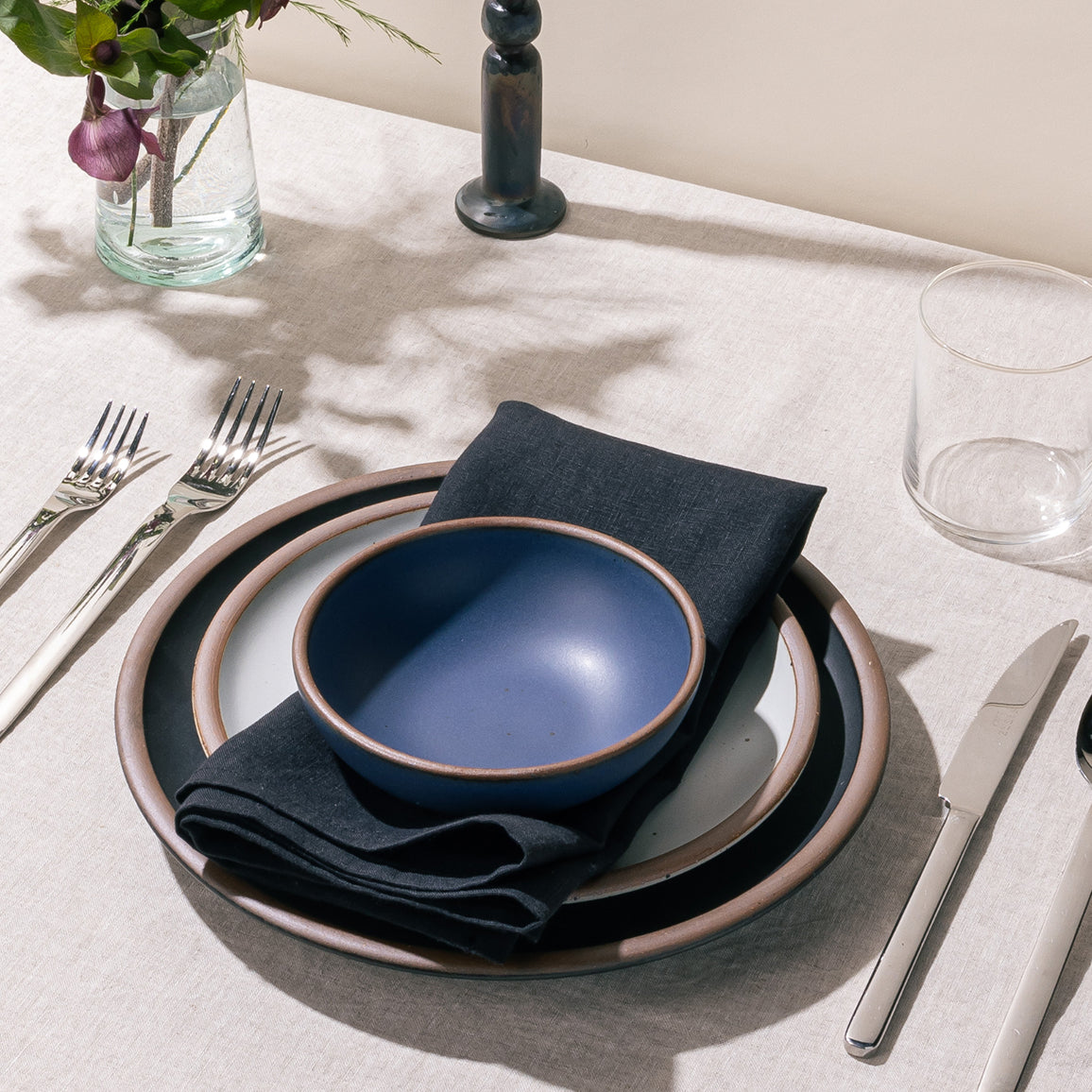 Blue ceramic bowl with unglazed rim on stacked plates and a dark linen napkin, styled with silverware, glassware, and flowers.