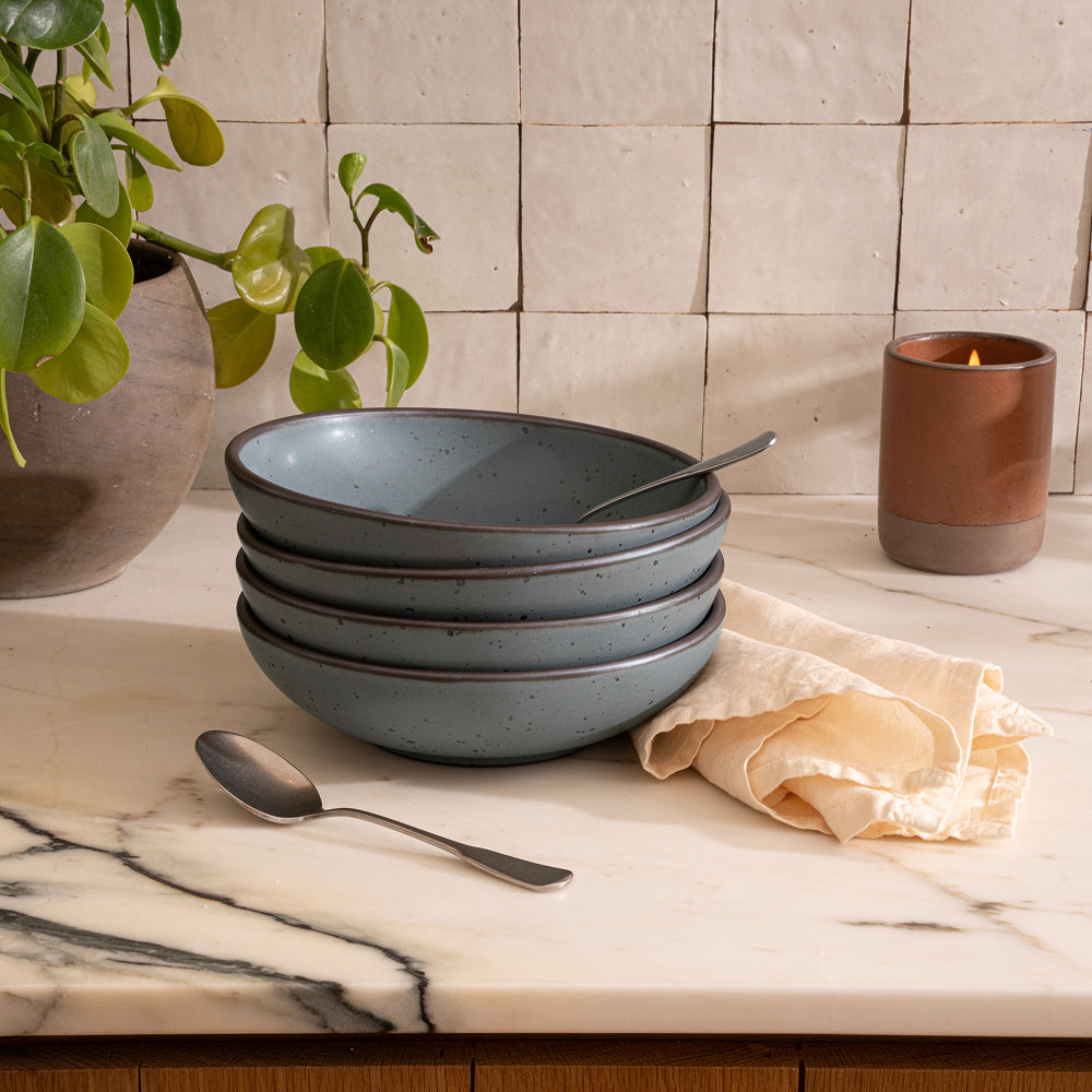 A stack of shallow ceramic bowls in a blue-grey color with silverware and a candle on a kitchen countertop.