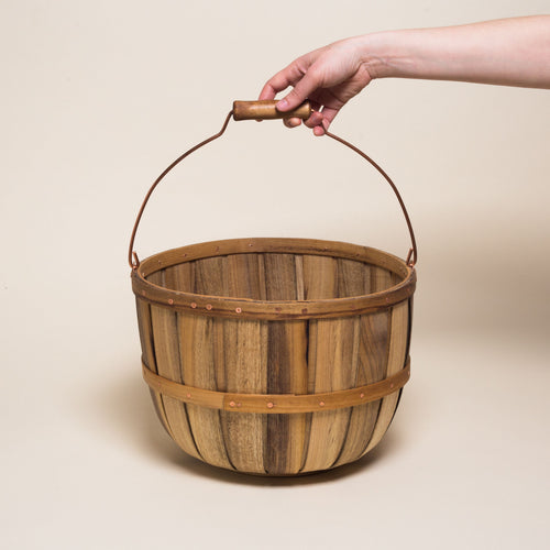 A wooden basket with metal handle and copper detailing is held by a hand against a plain beige background.