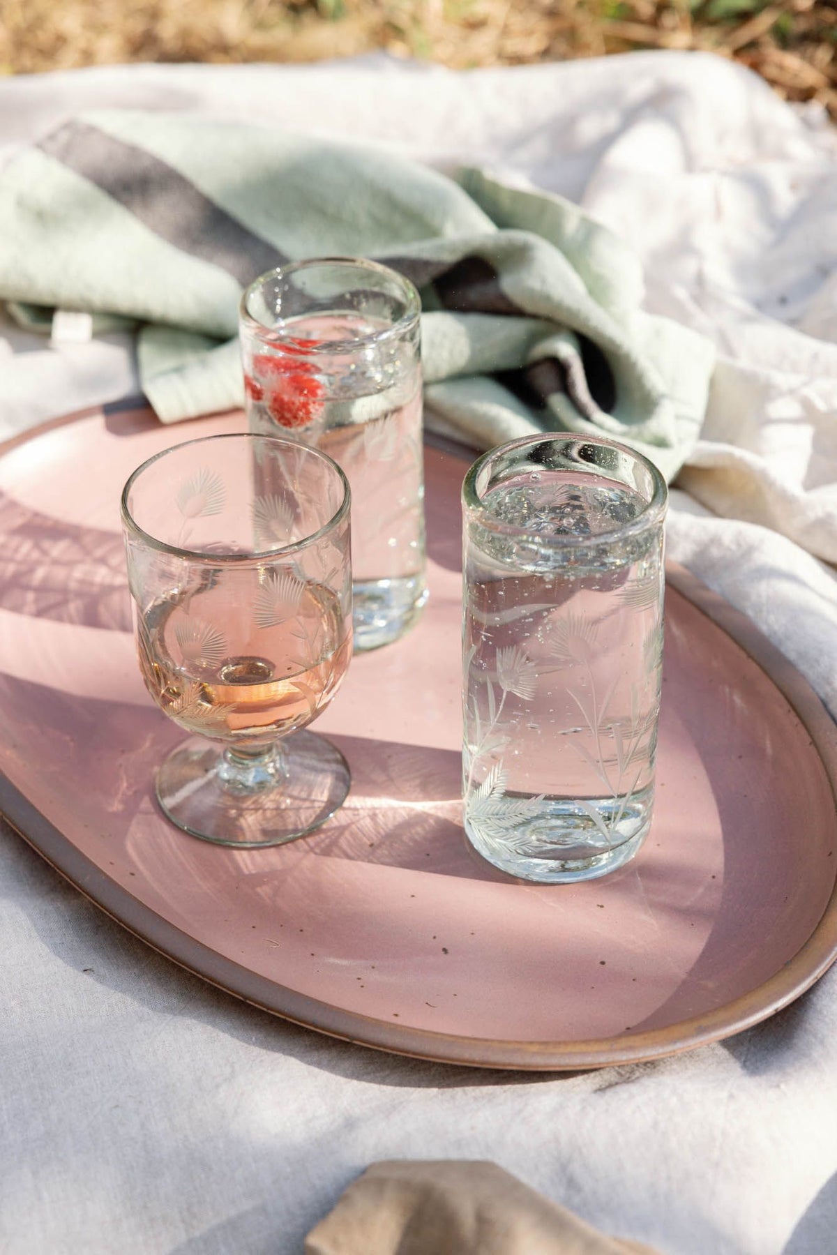 Three etched glass tumblers with beverages on a pink ceramic tray, set on a picnic blanket with a green and gray striped cloth in the background.