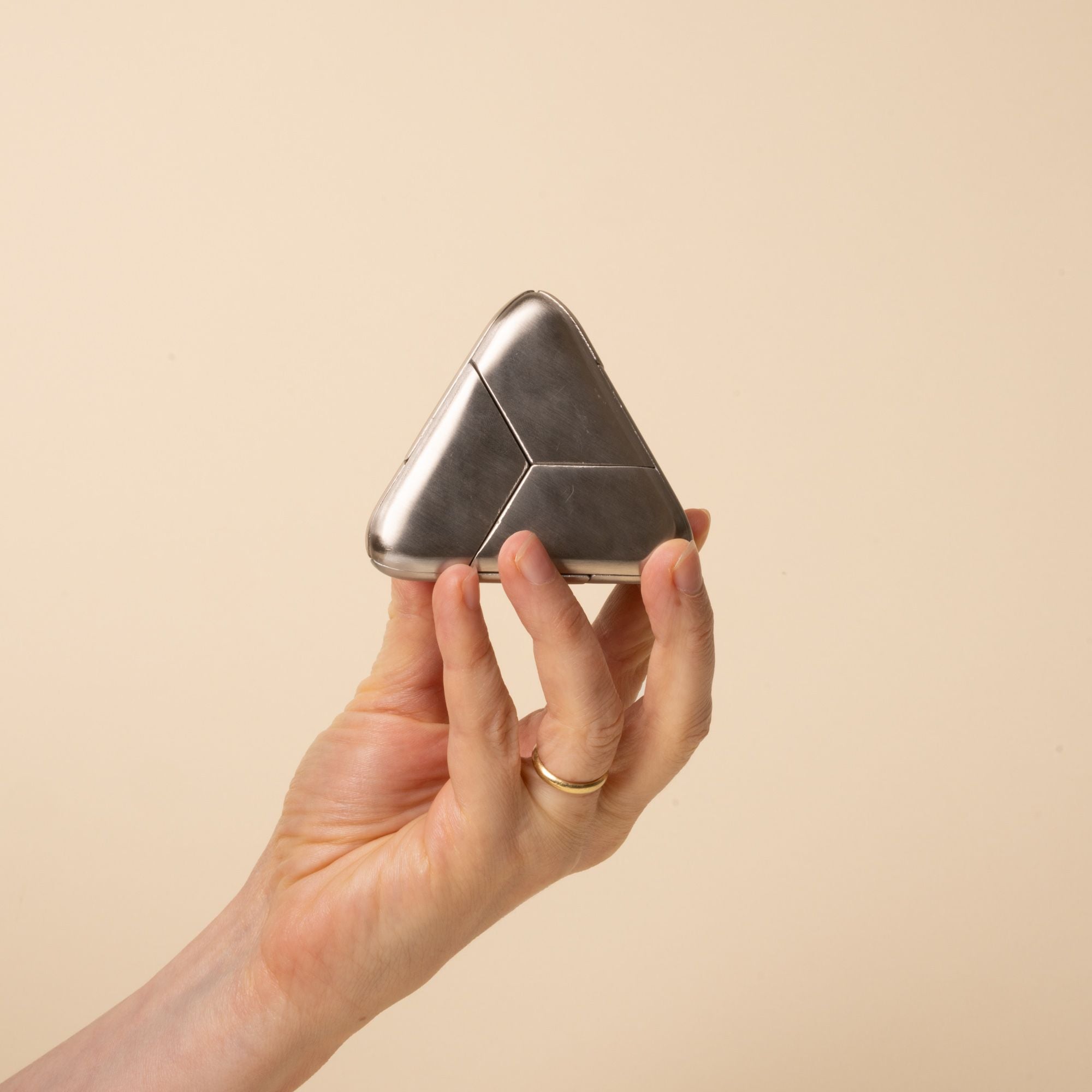 A hand holds a triangular silver metal puzzle with smooth rounded edges and subtle seam lines, resting on a beige background.