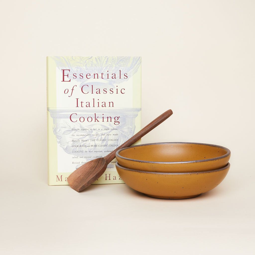 Cookbook titled "Essentials of Classic Italian Cooking" placed upright beside two stacked brown ceramic bowls with a wooden spatula resting on them, set against a neutral background.
