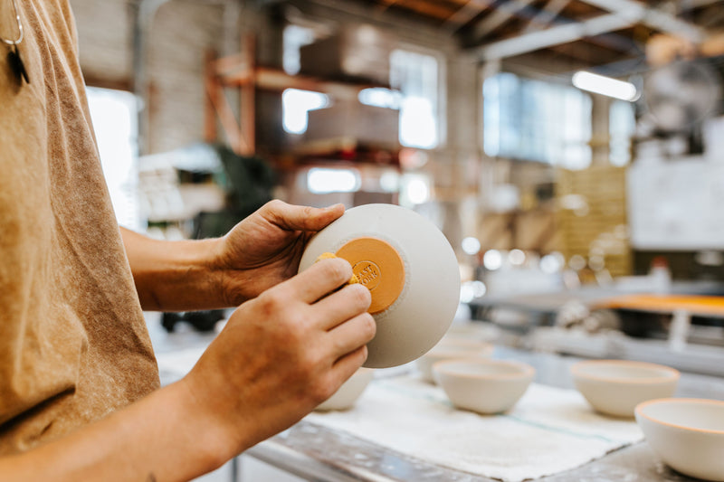 A person's hand removing glaze from the bottom of a ceramic bowl glazed in an off-white color, with a factory floor in the background.