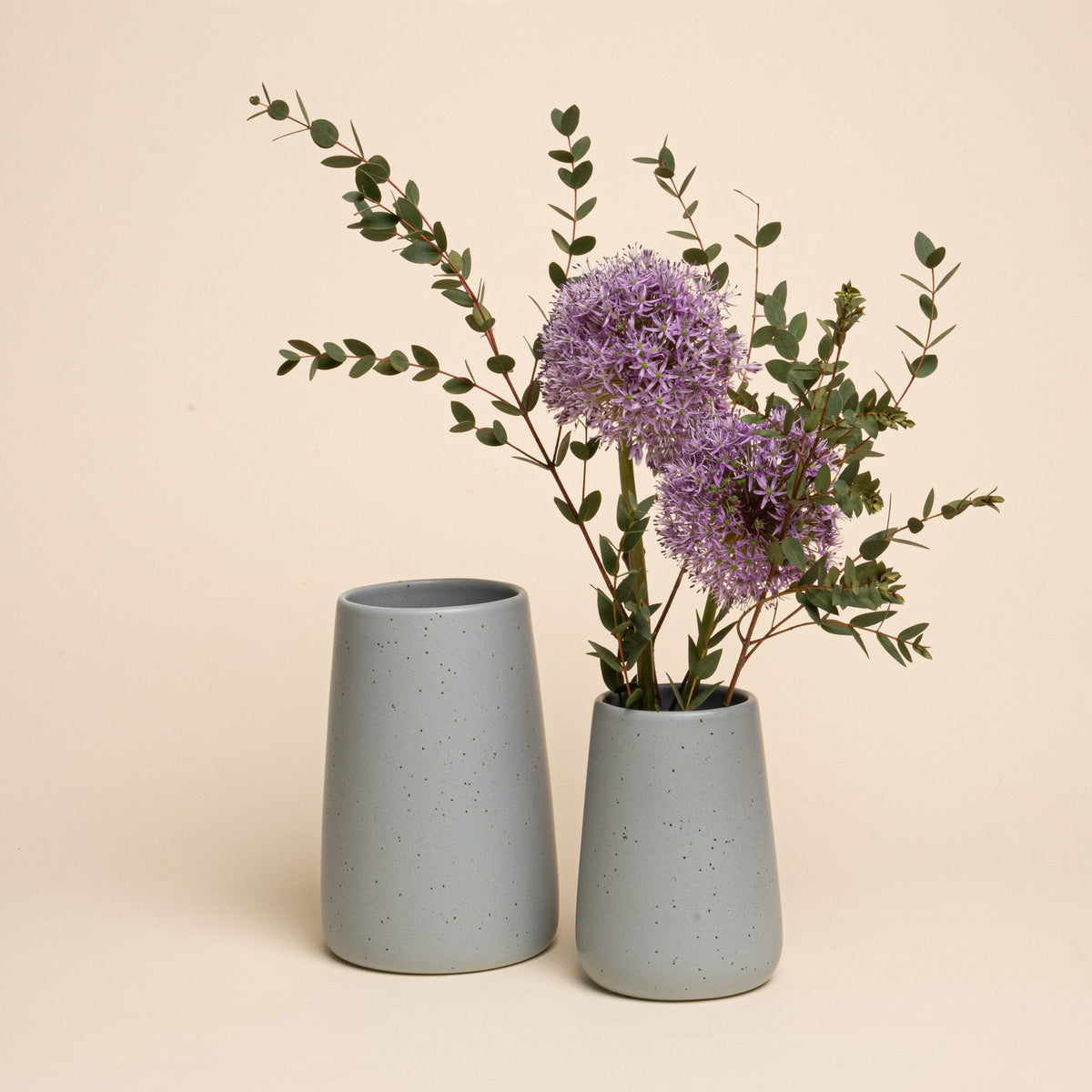 Two speckled blue-grey vases, one filled with purple flowers and greenery, styled on a neutral background.