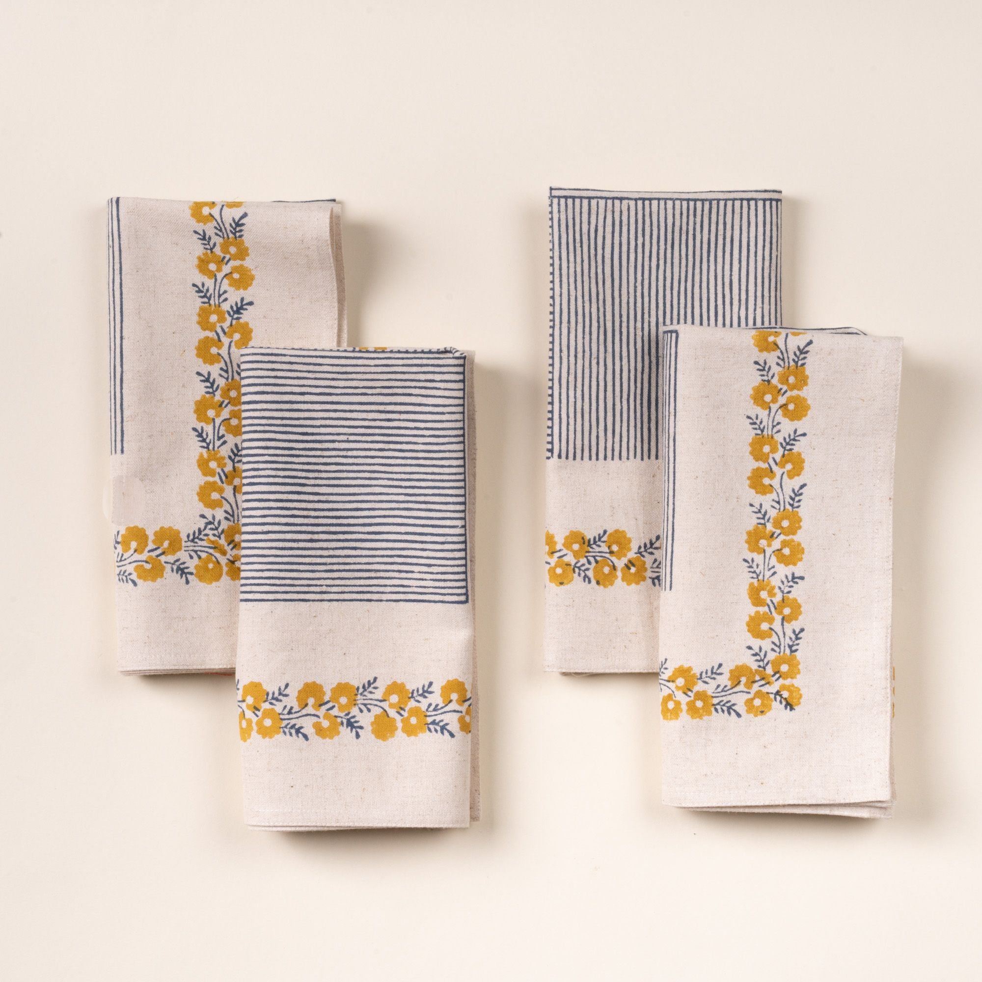 A set of four folded cream-colored napkins with blue striped accents and yellow floral border detailing, arranged neatly on a light background.