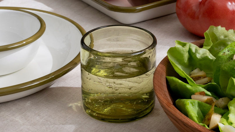 An olive green short transparent drinking glass, filled with water, on a tablescape with white enamel plates and bowls, and tomatoes and salad.