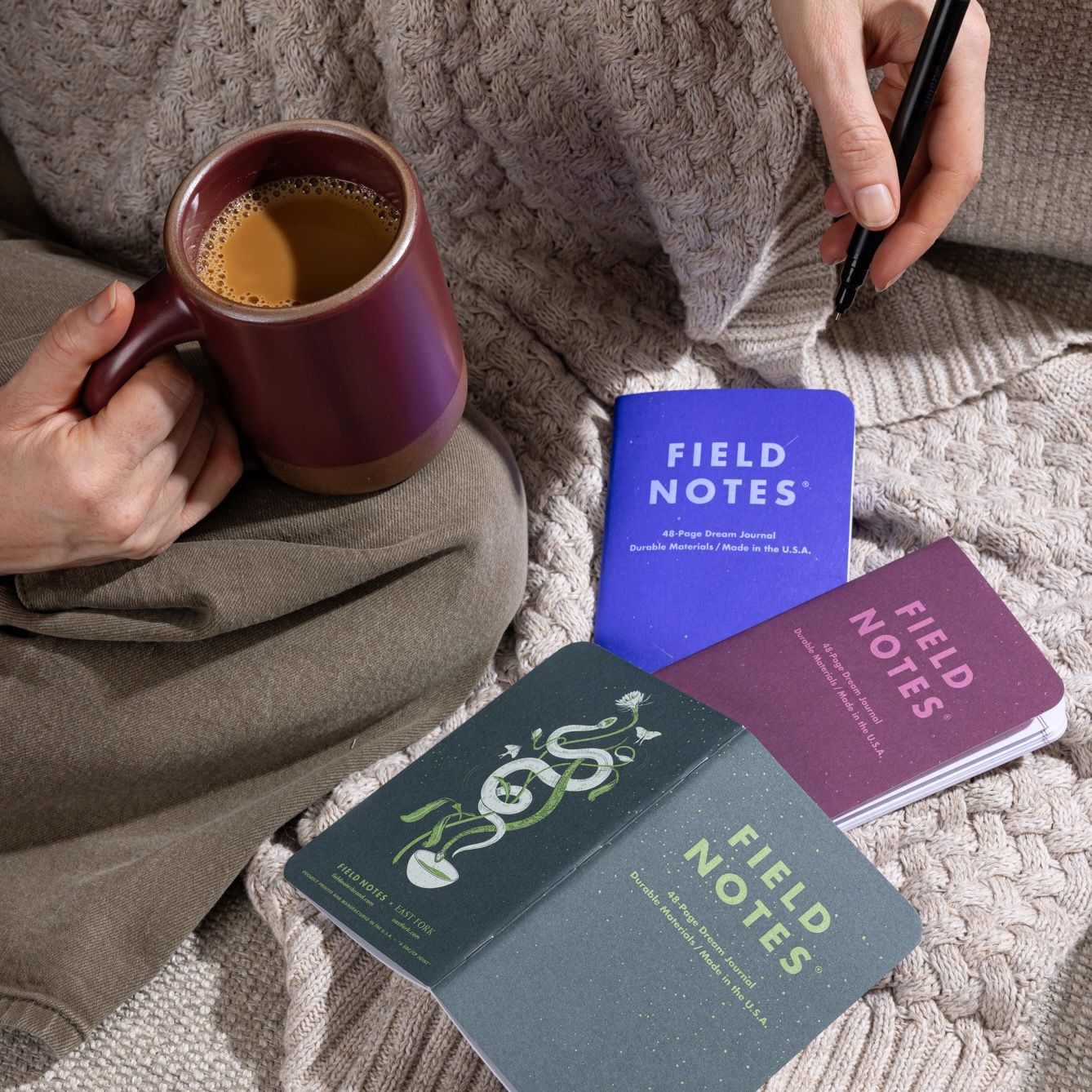 A person sits on a textured blanket holding a maroon ceramic mug filled with coffee, surrounded by three colorful Field Notes notebooks. They are writing with a pen, adding to the cozy and creative atmosphere.