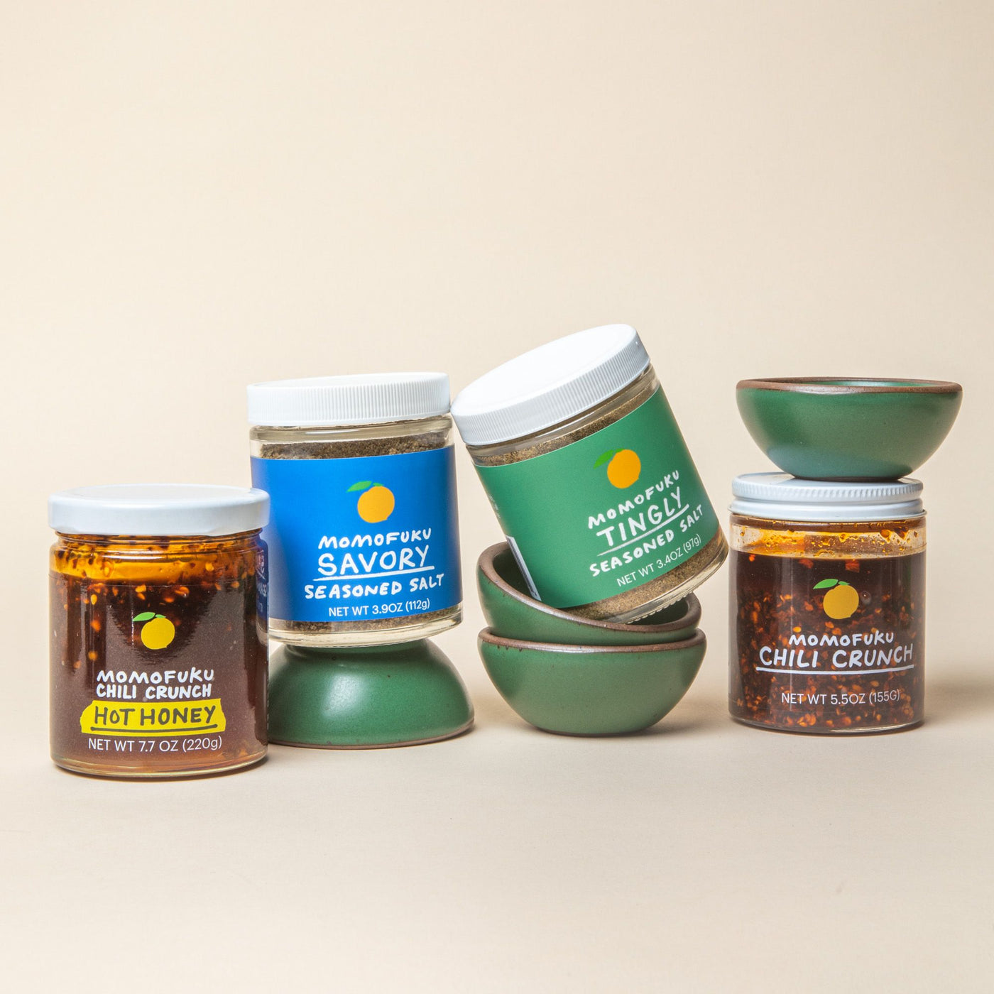 Assorted jars of Momofuku seasonings, including Savory and Tingly seasoned salts, and Chili Crunch, are displayed alongside green ceramic bowls on a beige background.