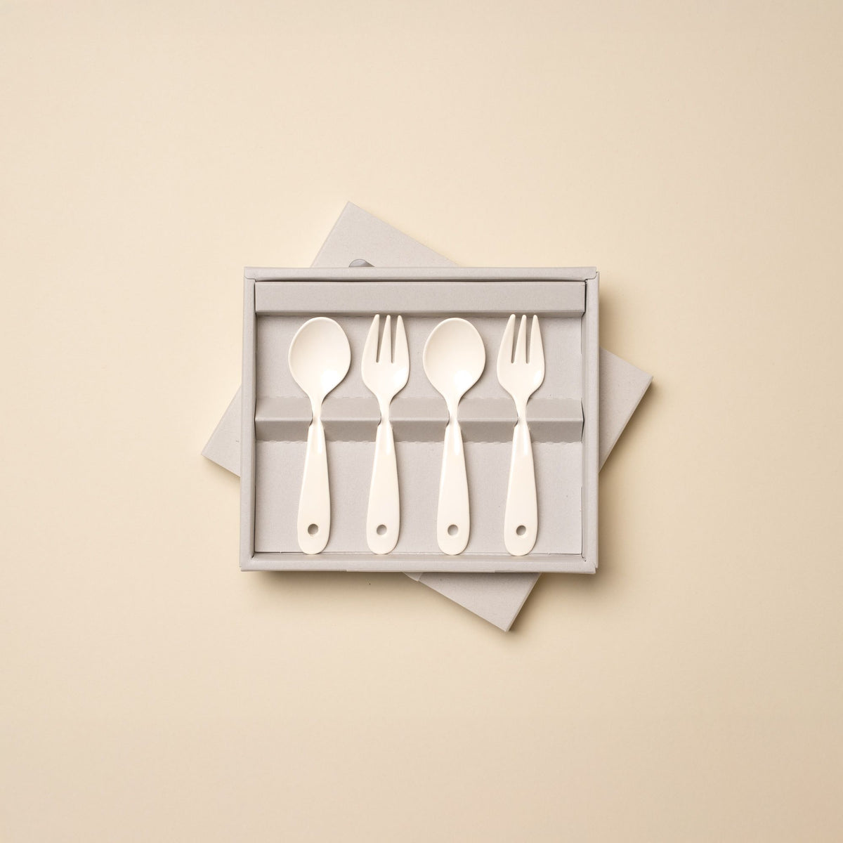 A boxed set of four white plastic toddler utensils, consisting of two spoons and two forks, displayed against a beige background.