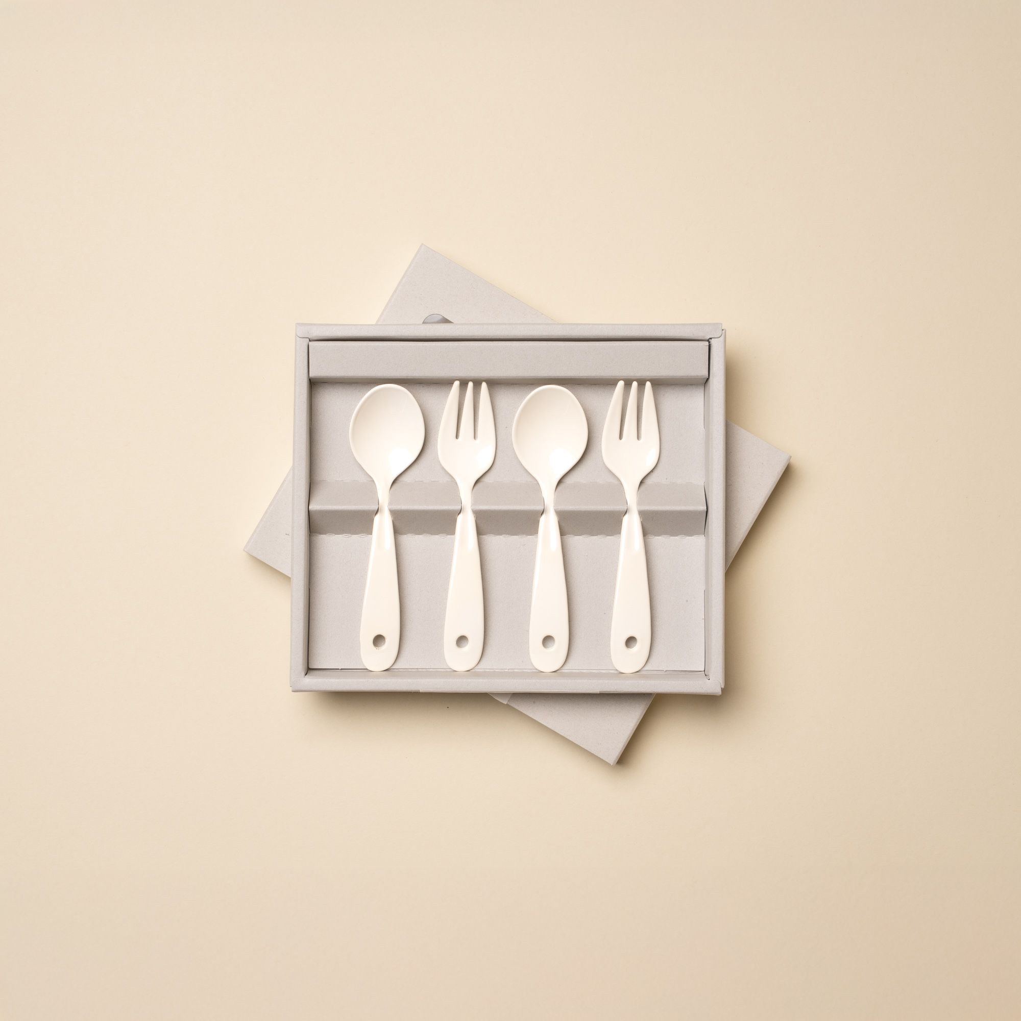 A boxed set of four white plastic toddler utensils, consisting of two spoons and two forks, displayed against a beige background.