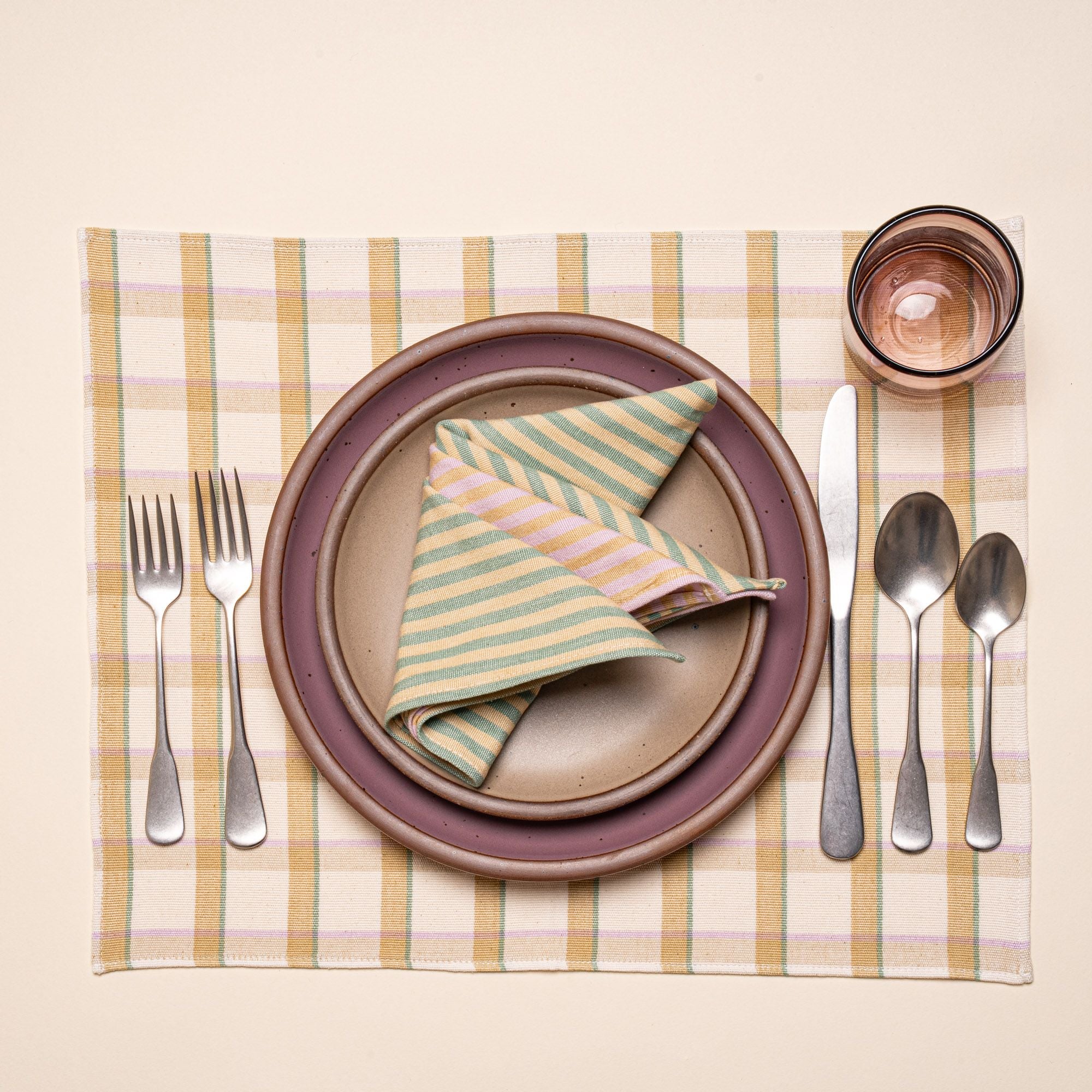 A place setting featuring ceramic plates in warm pale brown and soft mauve purple colors, a folded striped napkin, silverware, and a glass, on a pastel plaid placemat.