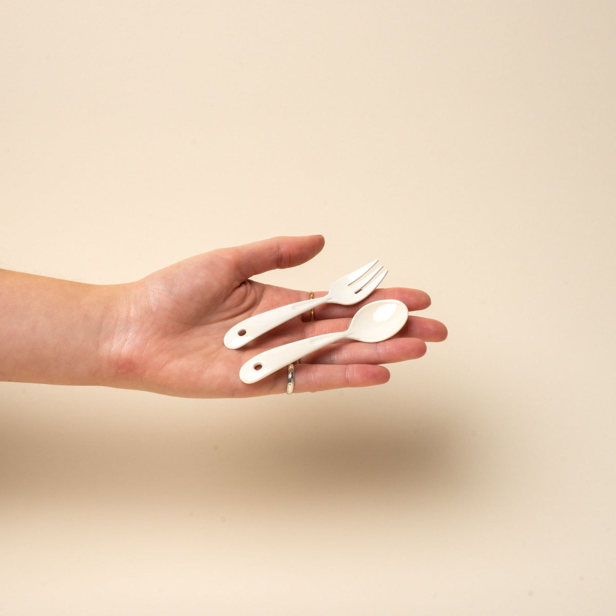 A hand holding 2 white enamel mini spoon and fork.